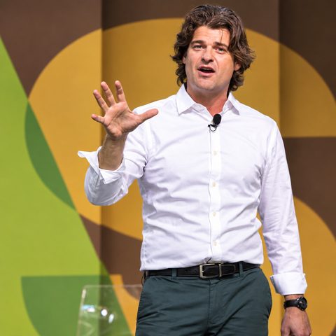 Dr. Brian Hare speaking on stage in a white shirt, gesturing with an open hand in front of a colorful geometric backdrop